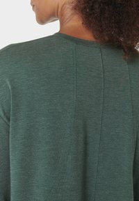 Vrouw met krullend haar die een donkergroen shirt draagt, gezien van achteren, met de focus op de textuur van de stof en de naaddetails van het shirt.