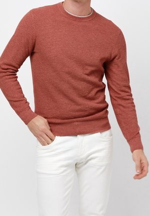 Homme portant un pull en tricot couleur rouille et un pantalon blanc, debout avec une main dans la poche sur un fond uni.