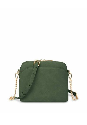 Woman in white coat and knit hat smiles, holding green handbag outdoors near snow-covered pine trees in winter setting.