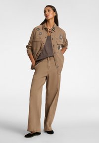 Vaaleanruskea farkkutakki, jossa on applikaatioita, päällä harmaa paita; yhteensopivat vaaleanruskeat leveät housut ja mustat tasapohjaiset kengät.