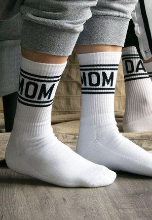 Twee paar benen met witte sokken met zwarte tekst, de ene zegt "MOM", de andere, gedeeltelijk zichtbaar, zegt "DAD", staand op een houten vloer.