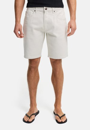 Mann iført lyse beige denimshorts med frontlommer og svarte flip-flops, stående mot en hvit bakgrunn.