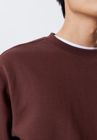 Brauner Sweatshirt mit Rundhalsausschnitt, glatter Textur und dezenten Nähten. Über einem weißen Hemd darunter getragen.
