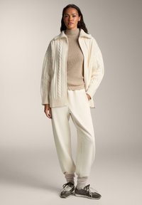 Cardigan oversized en tricot torsadé crème sur un col roulé beige, associé à un pantalon blanc ample et des baskets grises avec des accents blancs.