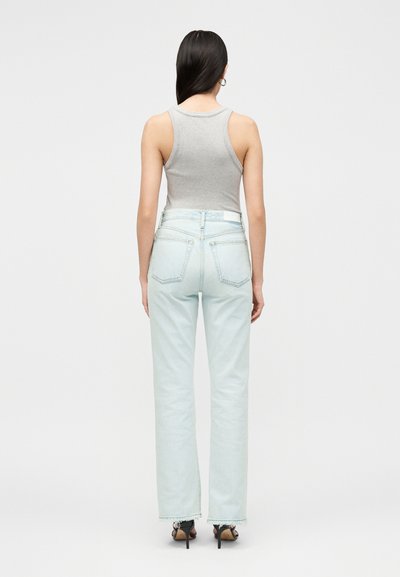 Femme aux longs cheveux foncés portant un top gris sans manches, un jean bleu clair taille haute et des sandales noires à talons, de dos.