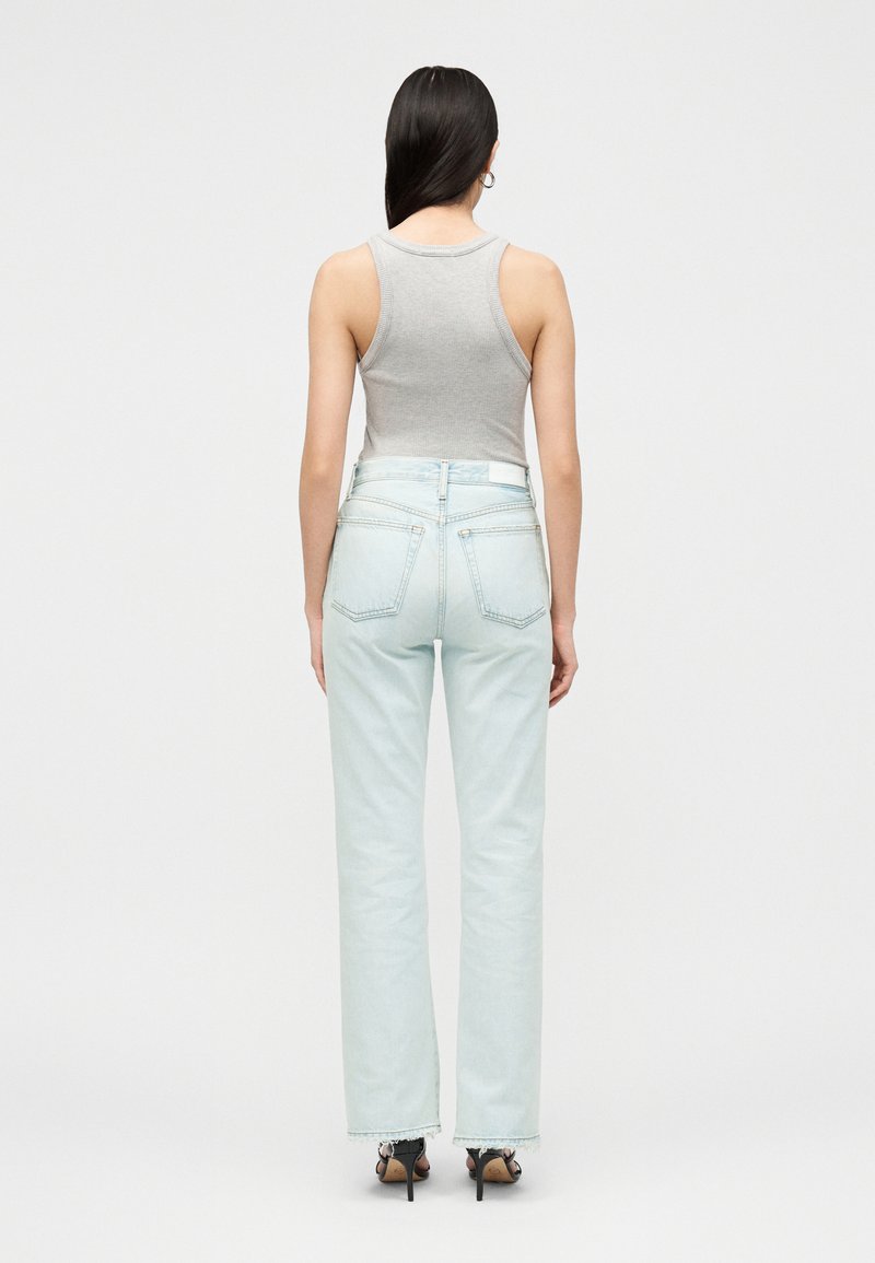 Femme aux longs cheveux foncés portant un top gris sans manches, un jean bleu clair taille haute et des sandales noires à talons, de dos.