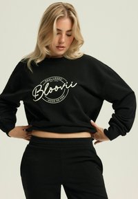 Czarna, oversize bluza z okrągłym białym wzorem z napisem "Bloovii" oraz "Real Looks Est. 2024". Miękki materiał, luźny krój, ściągacze przy mankietach.