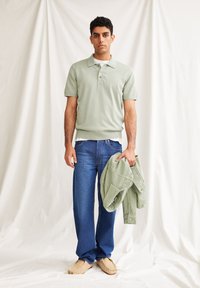 Lichtgroene poloshirt met een kraag, drie knopen, gecombineerd met een relaxed-fit blauwe spijkerbroek en beige instappers. Model houdt een groene jas vast.