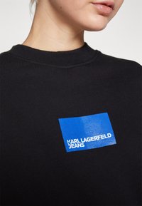 Sudadera negra con un logo rectangular azul brillante y texto blanco "KARL LAGERFELD JEANS" en el frente. Diseño clásico de cuello redondo.