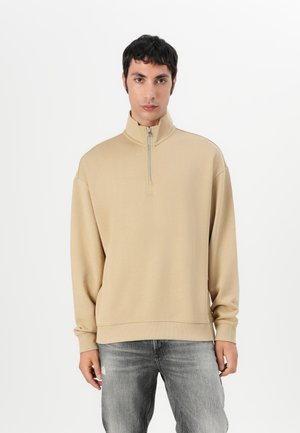 Sweatshirt beige à fermeture quart zippée avec un col haut et des poignets côtelés. Fabriqué en tissu doux, avec un détail de logo subtil sur la poitrine.
