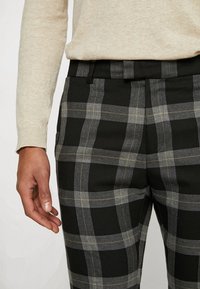 Osoba oblečená v černobílých kostičkovaných kalhotách a béžovém svetru s dlouhým rukávem, s levou rukou uvolněně položenou na boku.