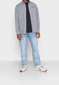 Světlomodrá košile na knoflíky s dlouhým rukávem a texturovanou látkou, tmavě šedé tričko pod ní, doplněné světle modrými džínami a šedými teniskami.