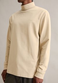 Beżowa koszula z długim rękawem i wysokim kołnierzem, wykonana z miękkiego materiału. Posiada subtelne logo w jasnym nadruku na piersi.