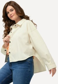 Crème-kleurige cropped jas met een puntige kraag, knoopsluitingen en lange mouwen. Draag het over een lichte top en combineer het met blauwe jeans.