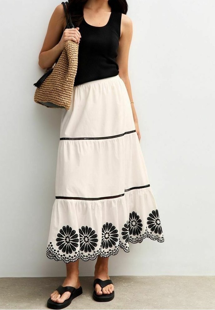 Femme portant un haut noir sans manches, une jupe blanche à volants avec broderie florale noire, des sandales noires et portant un sac tressé beige à l'épaule.