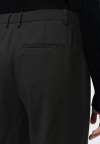 Prim plan cu pantaloni negri croiți pe comandă, cu buzunar spate, purtați cu un pulover negru cu nervuri, arătând șoldul și mâna dreaptă.