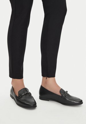 Person trägt schwarze enge Hosen mit Reißverschlüssen am Knöchel und schwarze Lederloafers mit Metall-Spangen-Detail vor weißem Hintergrund.