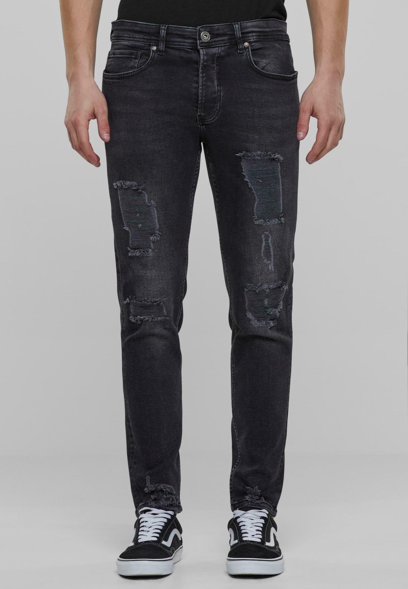 Vaqueros negros desgastados con detalles rasgados, corte ajustado, cinco bolsillos, cierre de botón de metal y un leve desvanecimiento. Combinados con zapatillas en negro y blanco.