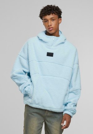 Light blue fleece pullover with half-zip, featuring a black patch at the chest, elastic cuffs, and a relaxed fit.