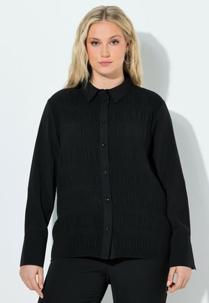 Mujer rubia con pendientes de aro plateados y una camisa negra abotonada con pliegues horizontales texturizados, posando frente a un fondo claro y sencillo.