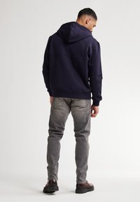 Sudadera con capucha azul marino de textura suave, combinada con jeans ajustados gris claro y botas marrón oscuro. Vista trasera.