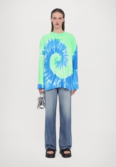 Bright green and blue tie-dye long-sleeve shirt with a swirl pattern, paired with faded blue wide-leg jeans and black platform sandals. Silver handbag.