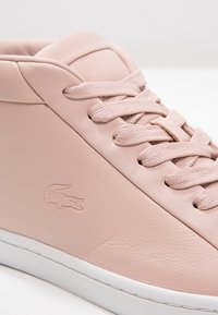 Gros plan d'une basket en cuir rose avec des lacets assortis et une semelle blanche, avec un logo crocodile embossé sur le côté.