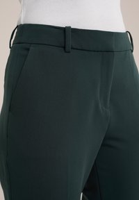 Mörkgröna skräddarsydda byxor med en slät yta, snedställda fickor och en smal passform. Innehåller bälteshällor och en framköt för struktur.