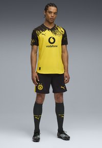 Hombre de pie con un uniforme de fútbol amarillo y negro del Borussia Dortmund que lleva los logos de Vodafone y Puma, con botas negras y medias a juego.
