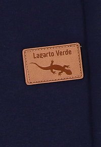 Bruin leren patch met de tekst "Lagarto Verde" en een silhouet van een hagedis, genaaid op een donkerblauwe stoffen achtergrond.
