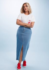 Valkoinen t-paita, jossa on punainen kirsikkagrafiikka, yhdistettynä siniseen farkkujen maksihameeseen, jossa on etuaukko, ja punaisiin lenkkareihin. Sileä pinta.