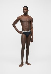 Αθλητικός άντρας που στέκεται ξυπόλητος με το ένα χέρι στη μέση, φοράει μαύρα εσώρουχα Nike με λευκή ζώνη με λογότυπο σε μονόχρωμο φόντο.