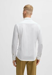 Chemise blanche à manches longues avec un col pointu, texture lisse et détails de plis au dos, portée avec un pantalon de couleur claire.