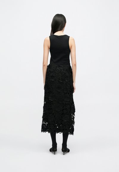 Fekete ujjatlan felső hosszú fekete szoknyával párosítva, amely részletes csipkés virágmintákkal és egyenes sziluettel rendelkezik, fekete lábbelivel viselve.