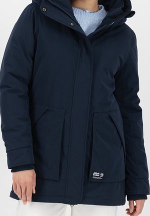 Personne portant une veste d'hiver à capuche bleu marine avec de grandes poches frontales et un pull en tricot bleu clair en dessous.