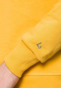 Żółta bluza z ribbowanymi mankietami i srebrnym logo na rękawie, charakteryzująca się miękką fakturą i luźnym krojem.