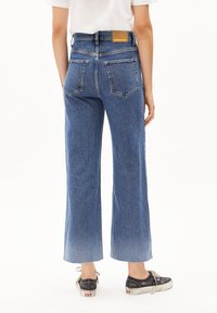 Jeans de mezclilla azul de pierna ancha con un lavado claro, que cuentan con una etiqueta de cuero en la cinturilla trasera y dos bolsillos traseros. Combinados con zapatillas negras.
