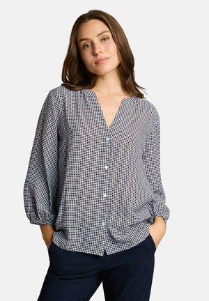 Femme portant un chemisier blanc à manches longues à pois noirs et un pantalon foncé, debout les mains dans les poches devant un fond uni.