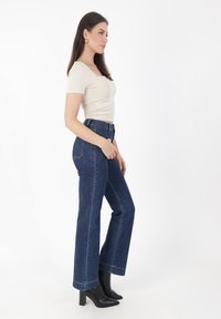 Top ajustado beige combinado con vaqueros acampanados de tiro alto en azul oscuro. El conjunto incluye botines negros con tacón y pendientes dorados sutiles.