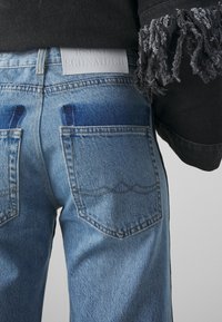 Vaaleansiniset farkut, joissa on haalistunut kuviointi, kaksi takataskua ja nahkainen etiketti. Mukana on musta teksturoitu vaate, jossa on fringet.