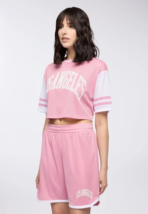 Giovane donna con indosso un completo sportivo Los Angeles in mesh rosa, top corto e pantaloncini, in piedi su uno sfondo chiaro e semplice.