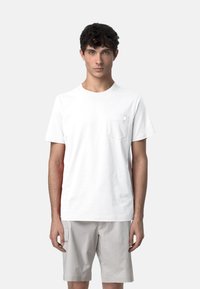 T-shirt bianca in cotone con collo rotondo, maniche corte e taschino frontale. Il modello indossa pantaloni corti grigi sotto.