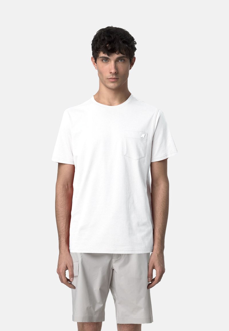 T-shirt bianca in cotone con collo rotondo, maniche corte e taschino frontale. Il modello indossa pantaloni corti grigi sotto.