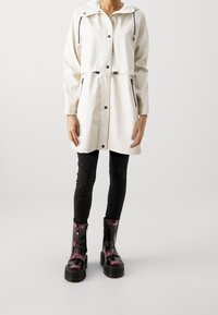 Hvid vandtæt jakke med hætte, sorte lynlåse og knapper, kombineret med sorte mønstrede leggings og blanke, mørkelilla ankelstøvler.