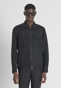 Giacca nera con zip, colletto, due tasche frontali e maniche lunghe. Il tessuto ha una texture liscia e una vestibilità sagomata. Indossata con occhiali da sole.