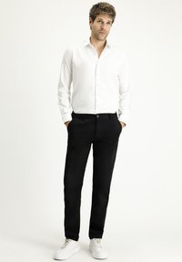 Mann mit hellbraunem Haar, der ein weißes Hemd mit Knöpfen, schwarze Hose und weiße Turnschuhe trägt und mit den Händen in den Taschen vor weißem Hintergrund steht.