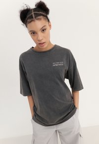 Graues Baumwoll-T-Shirt mit lockerer Passform, kurzen Ärmeln und rundem Ausschnitt; mit weißem Text "TAUCHE TIEFER IN DIE RUHE" auf der Brust.