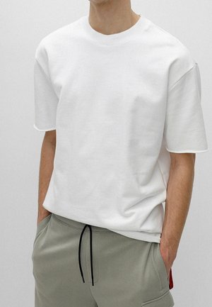 Mann trägt ein schlichtes weißes kurzärmeliges T-Shirt, leicht in hellgraue Jogginghose mit schwarzem Kordelzug gesteckt, Hände in den Taschen.