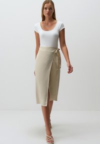 Gonna avvolgente beige con spacco laterale, legata in vita, abbinata a un top bianco aderente e sandali con lacci, su uno sfondo neutro.