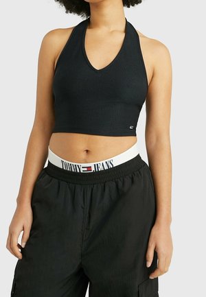 Vrouw draagt een zwarte geribde mouwloze crop top en zwarte broek, met een witte tailleband met het Tommy Jeans-logo.